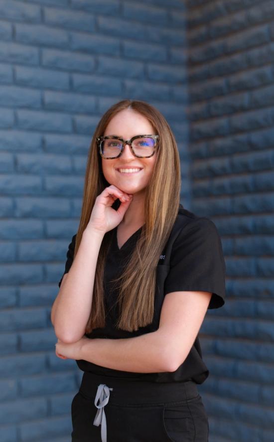 a woman wearing glasses and a scrub top stands in front of a brick wall