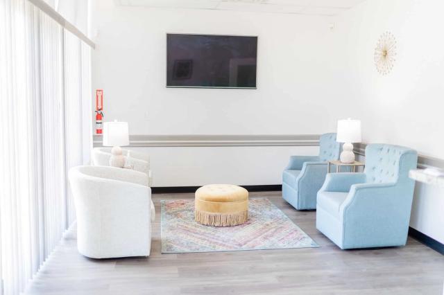 A bright waiting room with light blue and white armchairs, a yellow ottoman, a patterned rug, and a wall-mounted TV.