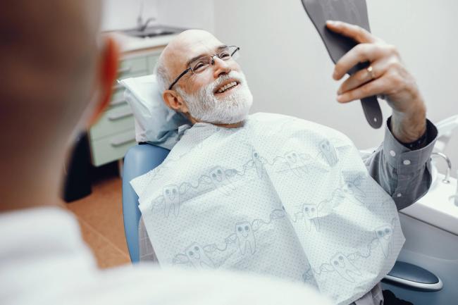 an elderly man is sitting in a dental chair looking at his teeth in a mirror .