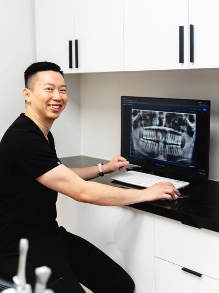 A smiling man in black scrubs looks at the camera while reviewing a dental X-ray on a computer screen.