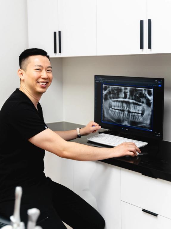 A smiling dentist in a black scrub top reviews a panoramic dental X-ray on a computer screen.