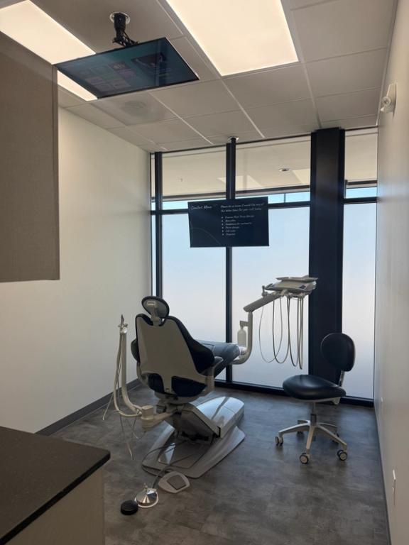 Modern dental exam room with a gray dental chair, instruments, frosted windows, and two screens.