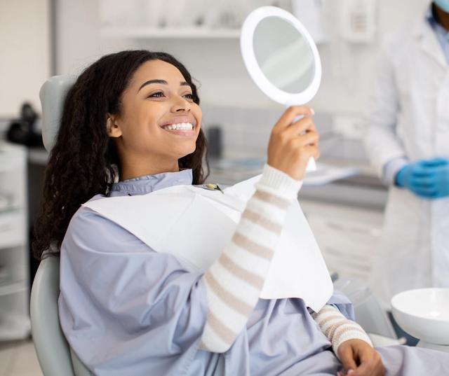 a woman is sitting in a dental chair looking at her teeth in a mirror .
