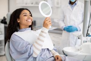 A smiling woman in a dental chair looks at her teeth in a handheld mirror, with a dentist in the background.