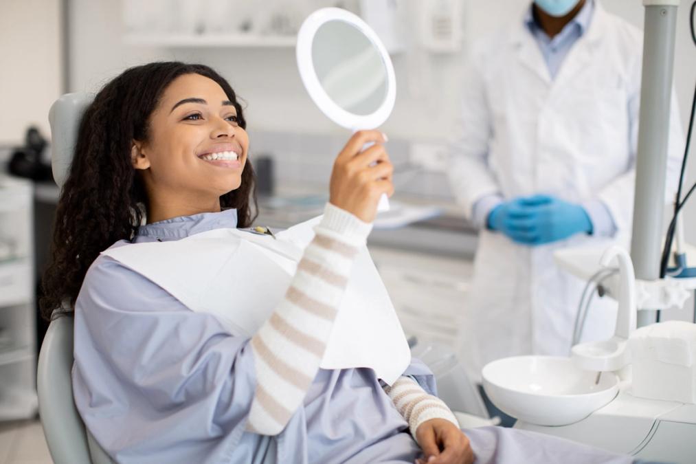 A smiling young Black woman in a dentist's chair looks at her reflection in a mirror.