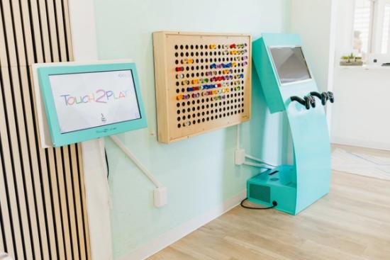 A light blue wall with three children's interactive games: a 'Touch2Play' touchscreen, a wooden pegboard with colorful pieces, and a turquoise arcade console.