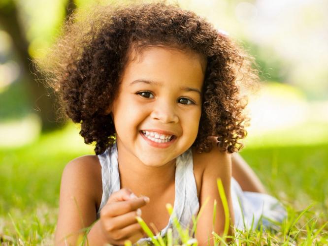 a little girl with curly hair is laying in the grass and smiling .
