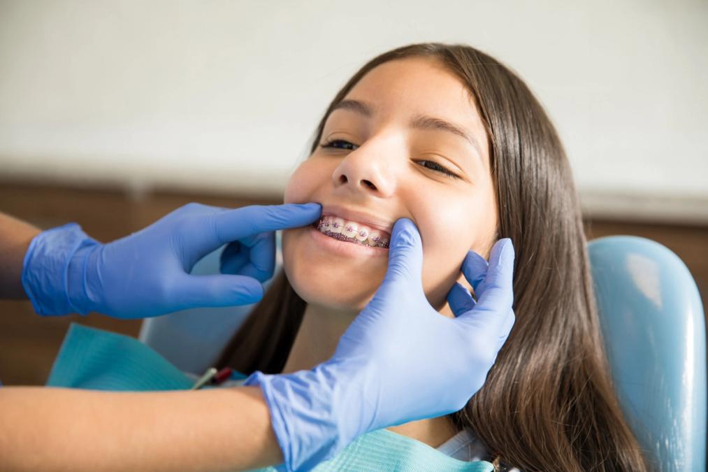A young girl with braces smiles as an orthodontist's gloved hands examine her teeth.