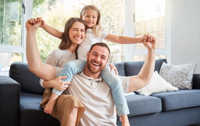 A joyful family of three, with a daughter on her father's shoulders and mother hugging him, all laughing in a living room.