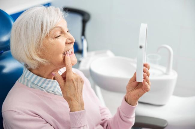 an elderly woman is sitting in a dental chair looking at her teeth in a mirror .