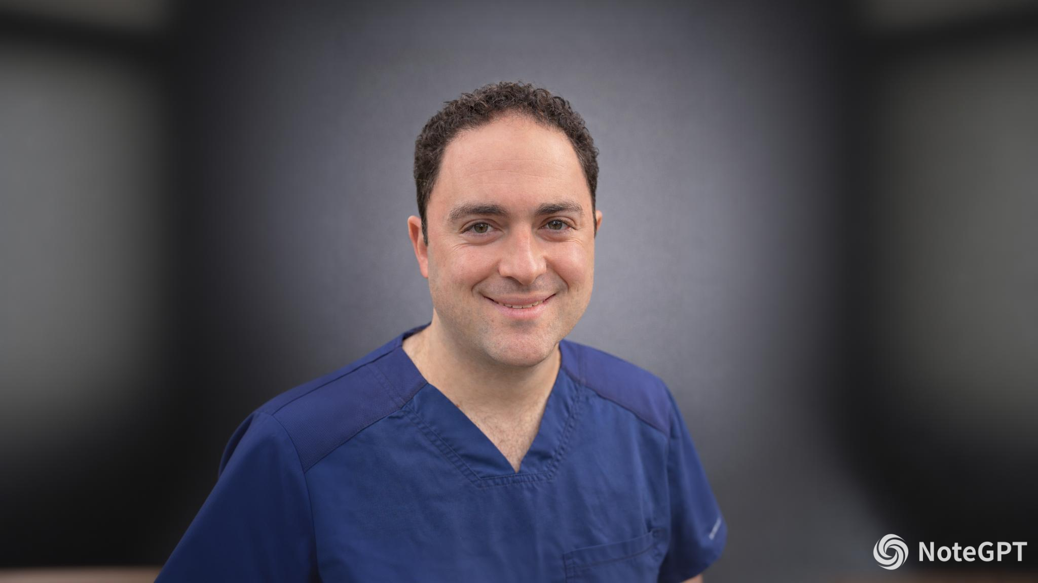 A smiling man with dark curly hair wears blue scrubs against a dark, blurred background.