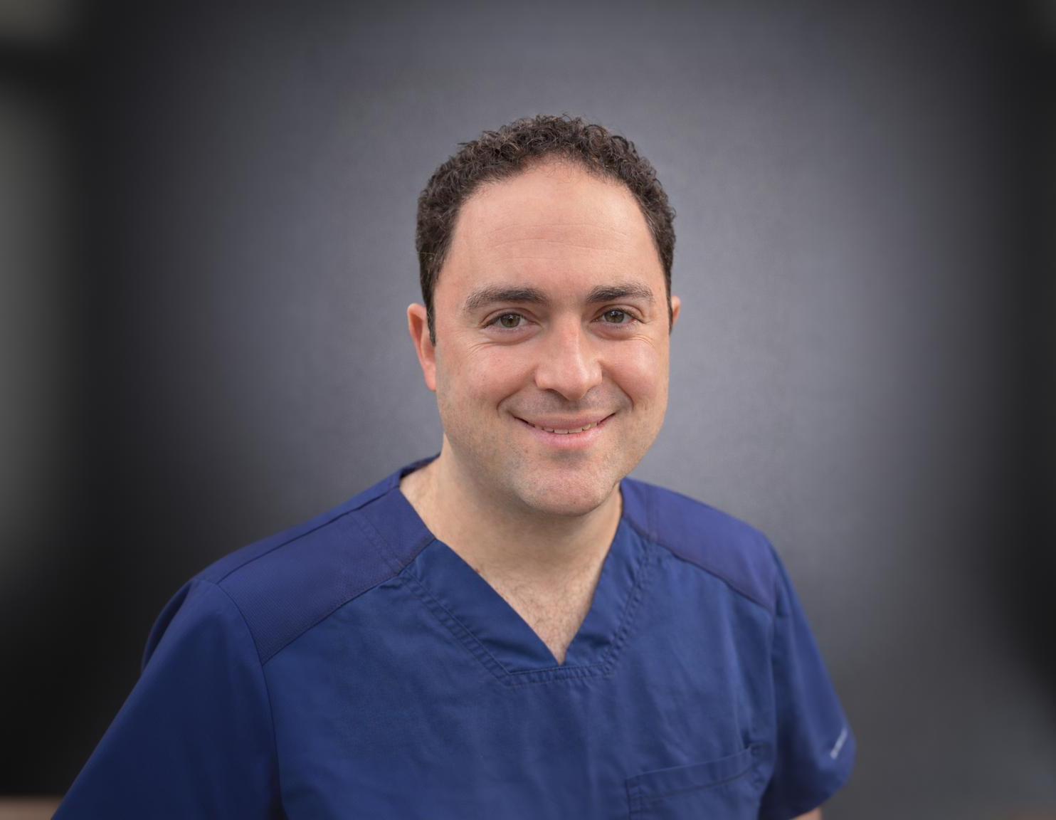 A man in blue scrubs smiling at the camera.