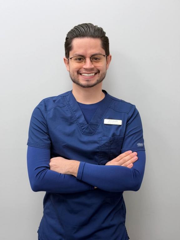 A smiling man in blue scrubs with a name tag reading JULIAN, standing with his arms crossed.