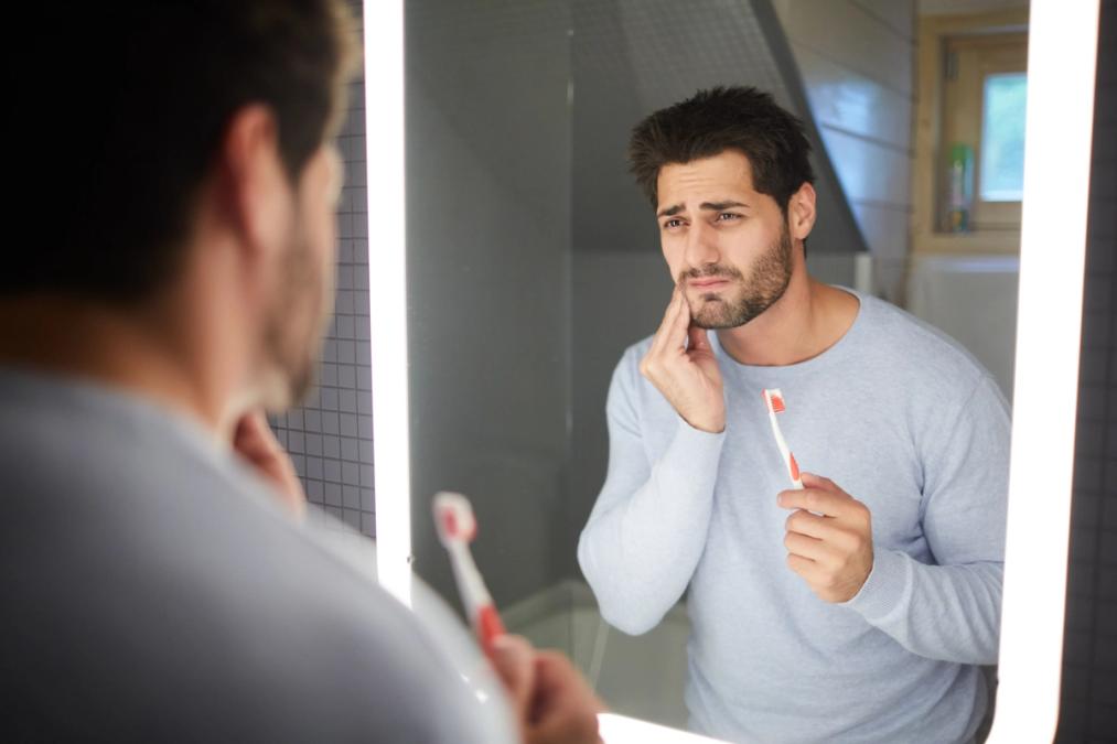 a man is brushing his teeth in front of a bathroom mirror and holding his face in pain.