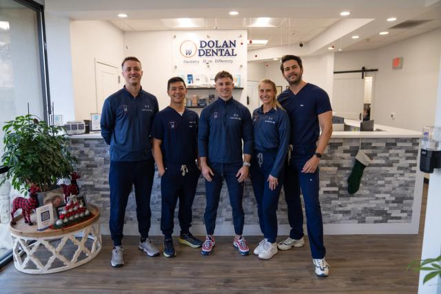 Five smiling dental team members in blue uniforms stand in the modern Dolan Dental office reception area, decorated for Christmas.