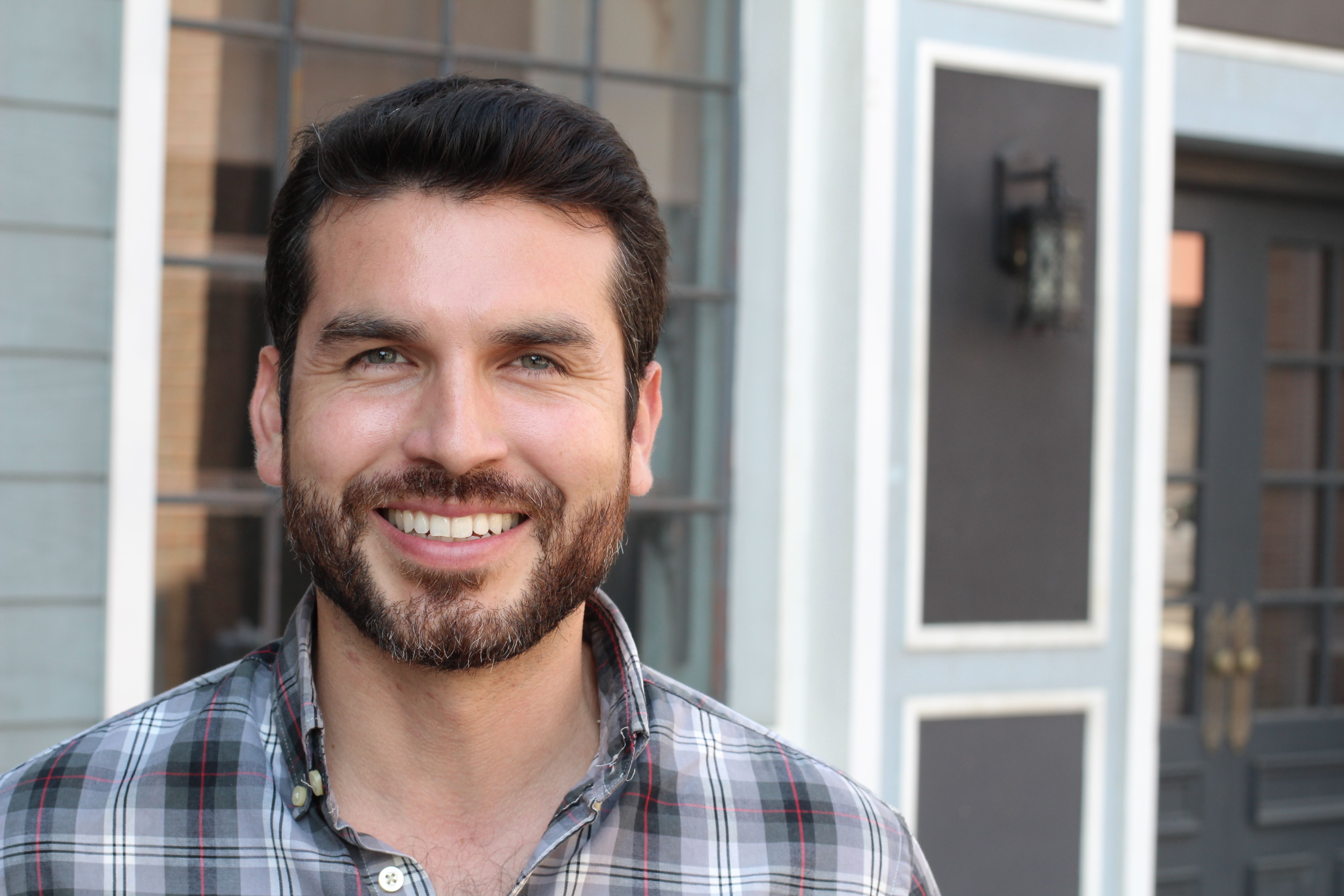 Smiling man with a beard in a plaid shirt outside a building.