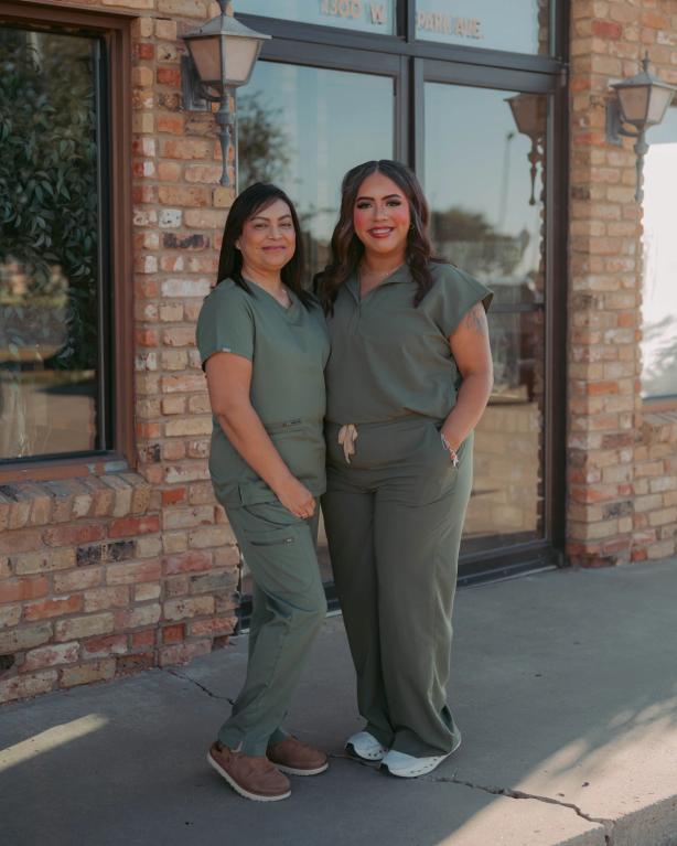 Two women in olive green scrubs smiling outside a brick building.