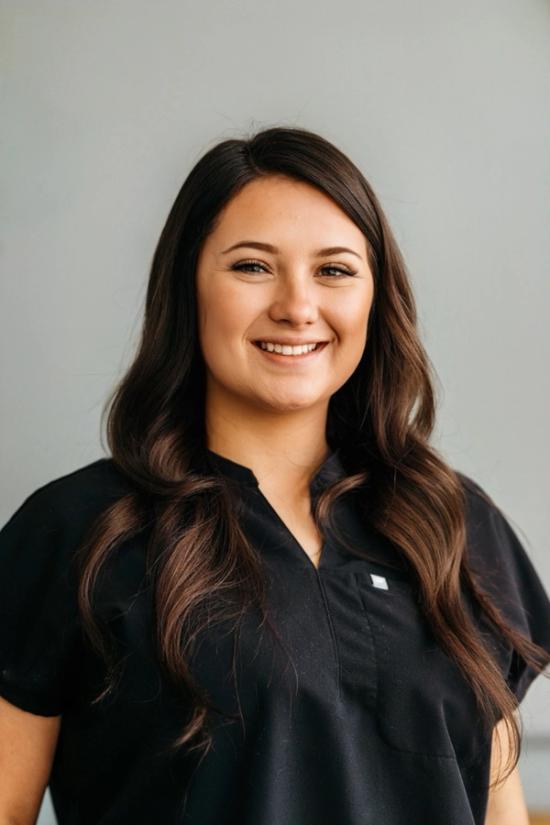 A smiling young woman with long brown hair wearing a black scrub top.