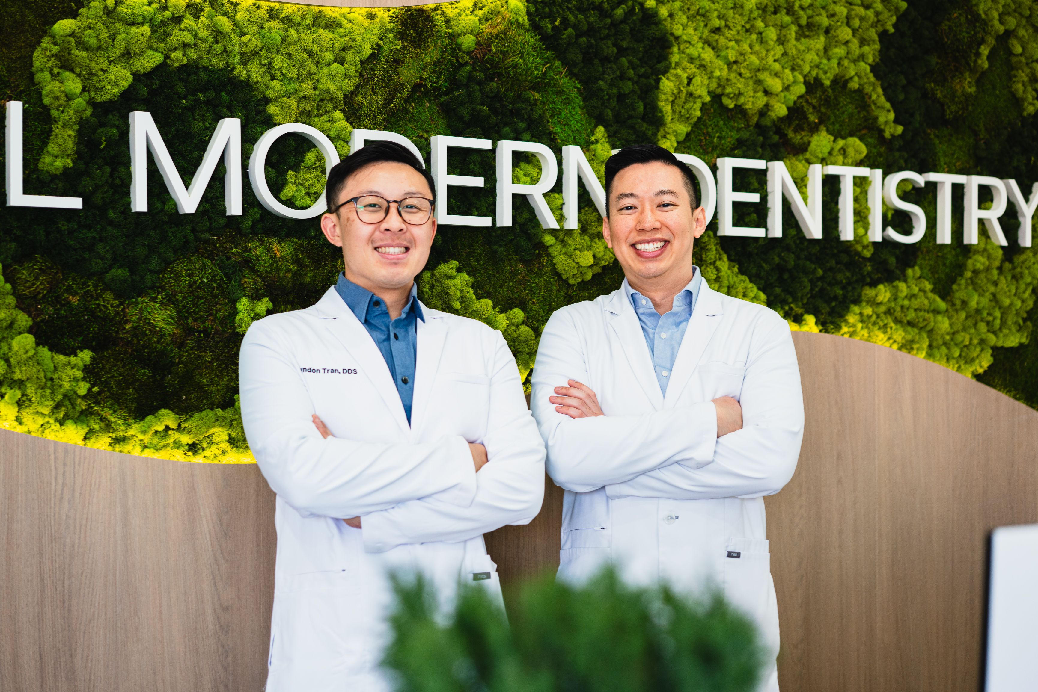 Two smiling male dentists in white lab coats stand with crossed arms in front of a green moss wall with "MODERN DENTISTRY" text.