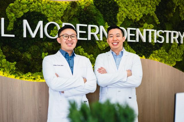 Two smiling male dentists in white lab coats stand in front of a green moss wall with "MODERN DENTISTRY" text.
