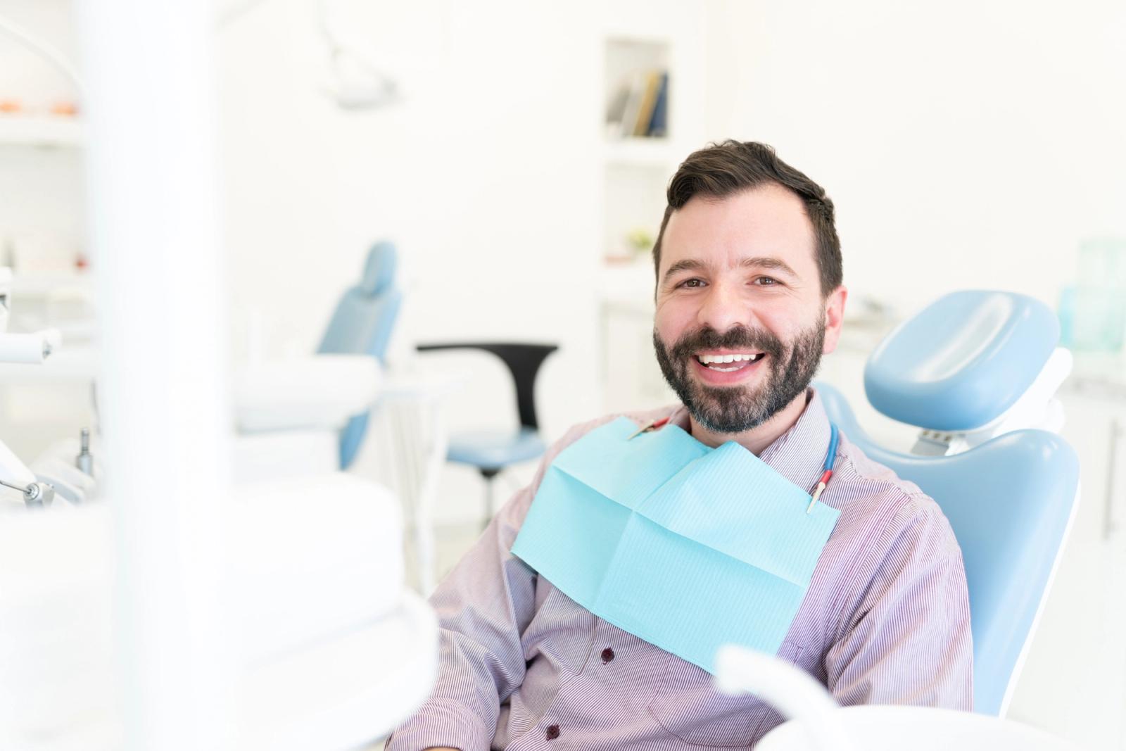 A smiling man with a beard sits in a dental chair with a blue bib.