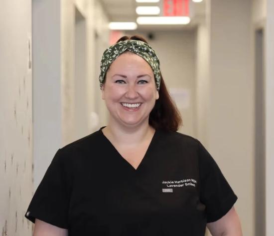 a woman in a black scrub top and headband is smiling in a hallway .