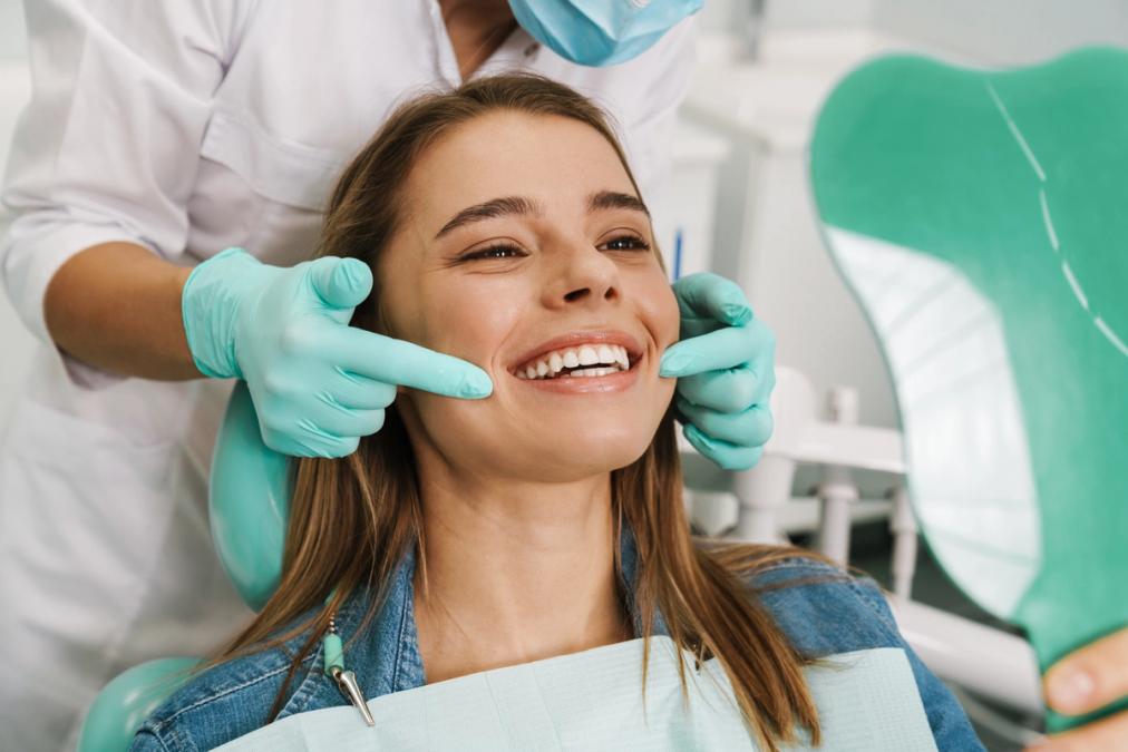 A smiling woman in a dental chair looks at her teeth in a mirror, while a gloved hand points to her mouth.