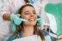 A smiling woman in a dental chair looks at her teeth in a mirror, while a gloved hand points to her mouth.