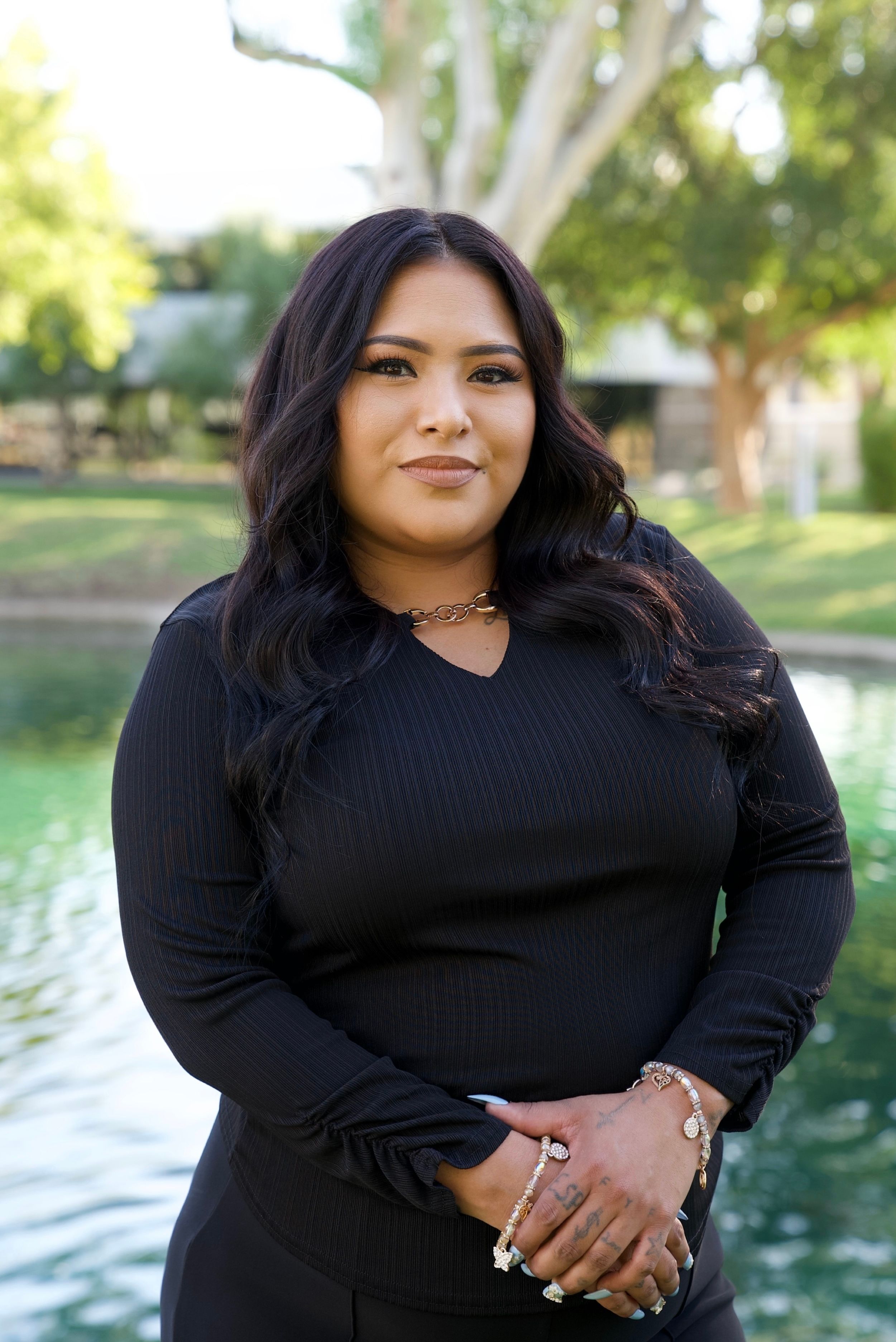 A woman with dark hair and a black top stands smiling by a body of water with trees in the background.