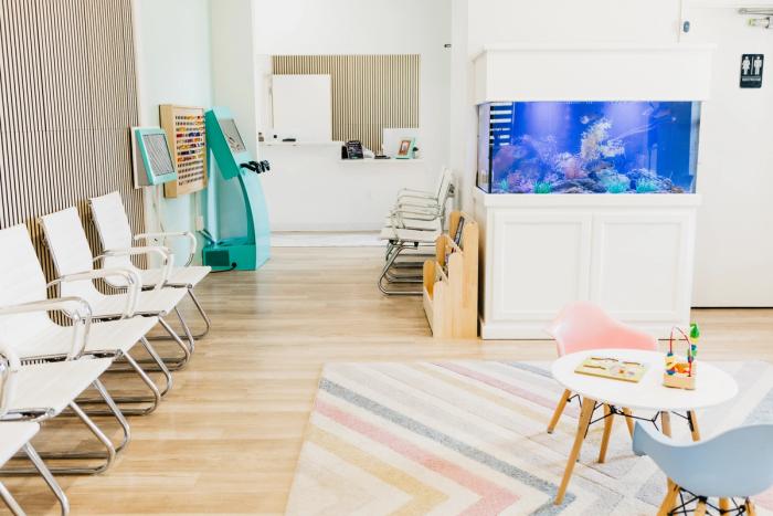 A bright, modern waiting room with white chairs, a large aquarium, and a colorful children's play area.