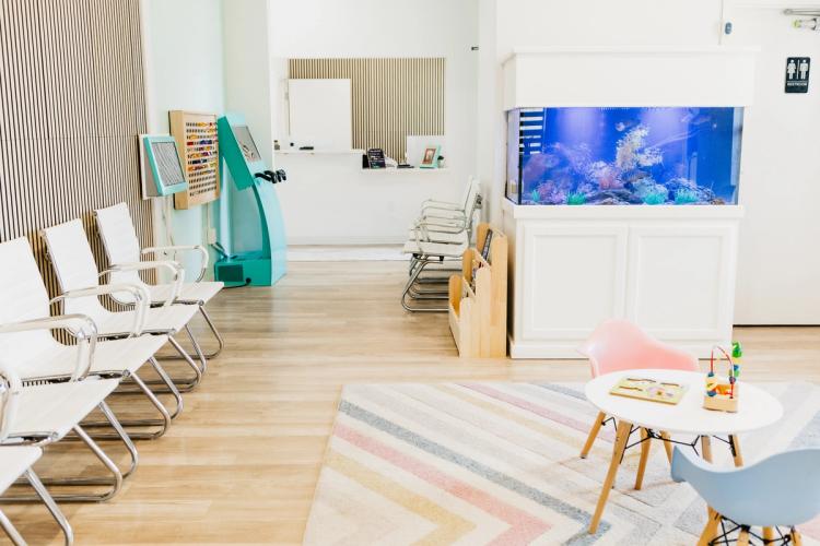 A bright, modern waiting room with white chairs, a large aquarium, and a colorful children's play area.