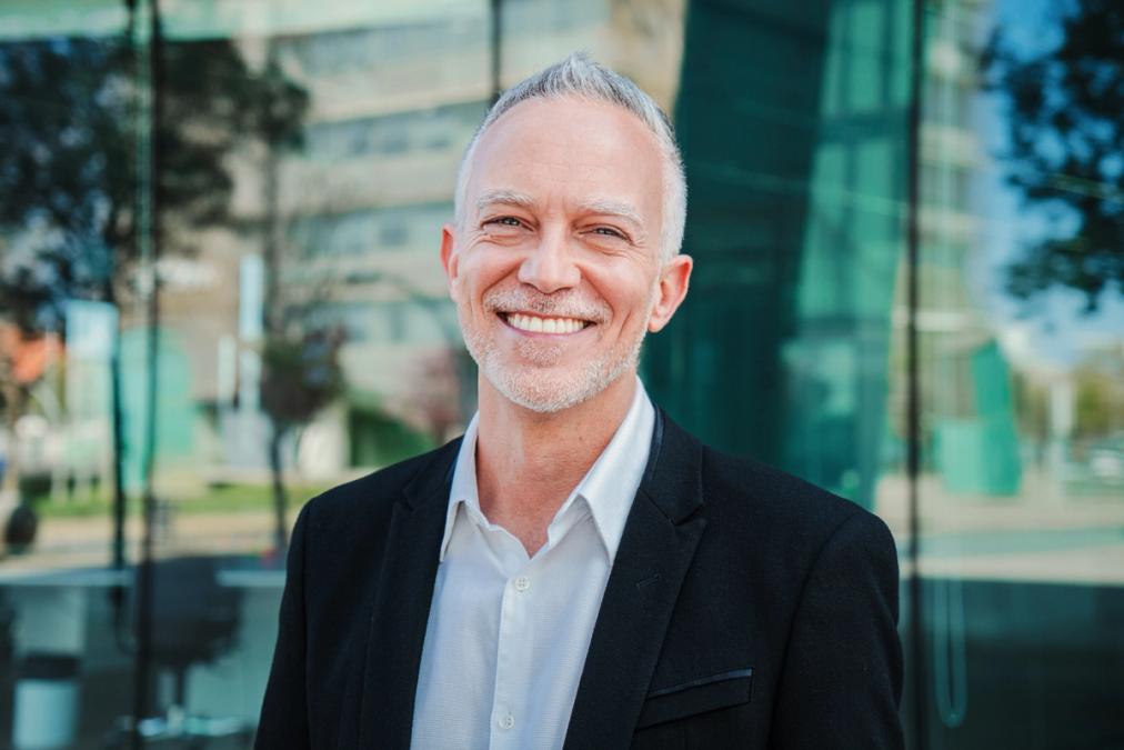 A smiling man with grey hair and a short beard in a black blazer, standing in front of a modern glass building.