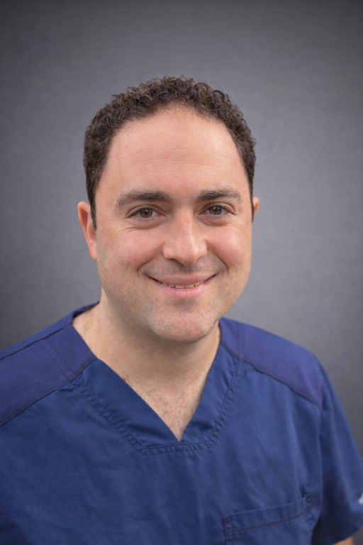 A smiling man with curly brown hair wears a blue scrub top against a gray background.