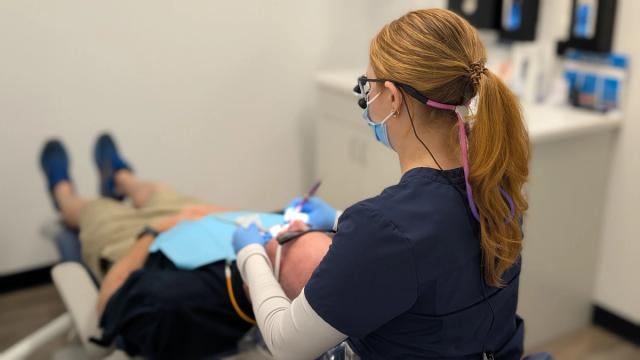 A dental professional wearing a mask and magnifying glasses works on a patient in a dental chair.