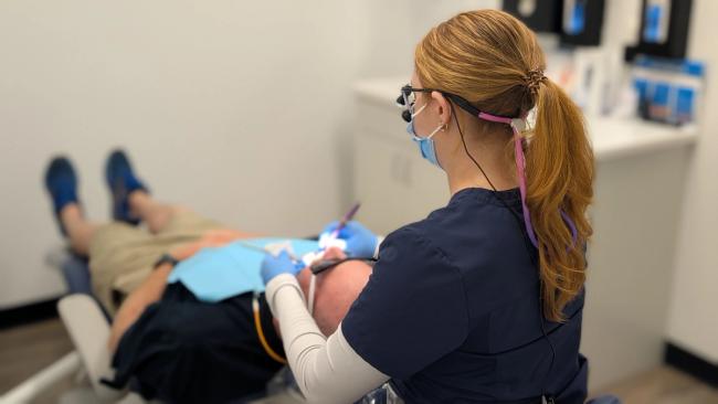 A dental professional wearing loupes and a mask works on a patient in a dental chair.