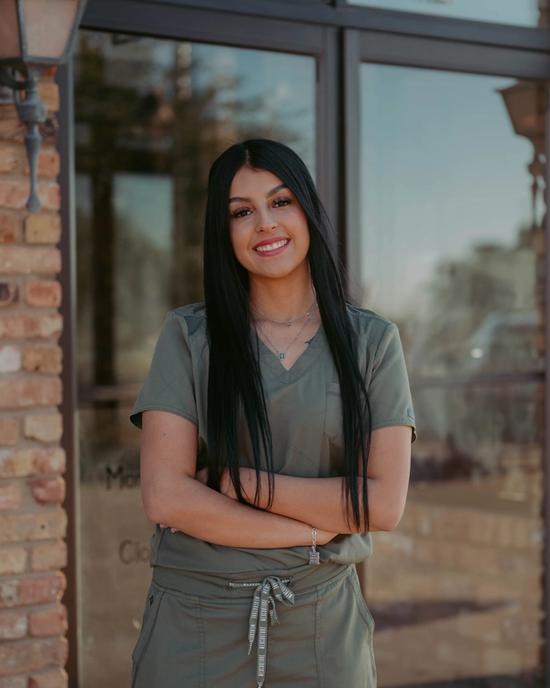 A smiling woman with long dark hair wearing olive green scrubs and crossing her arms.