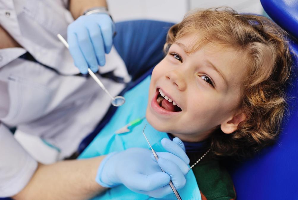 A laughing child in a dentist's chair, with a gloved dentist holding dental tools.