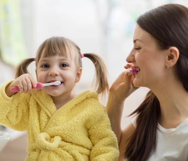 A smiling young girl in a yellow robe brushes her teeth next to her mother, who is also brushing her teeth.