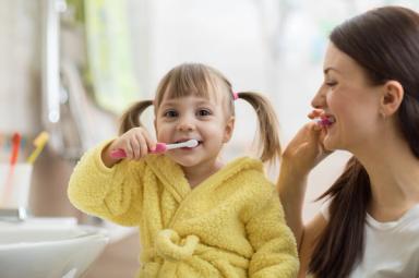 A happy young girl in a yellow bathrobe brushing her teeth with her mother.