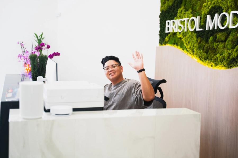 A person smiling and waving from behind a reception desk with a "BRISTOL MOD" moss wall sign in the background.