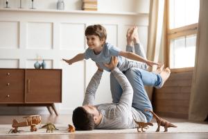 A father lies on his back, holding his smiling son aloft with his feet and hands as the boy pretends to fly.