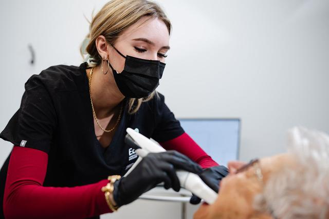 A clinician in a mask and gloves uses a handheld device to scan an older patient's face.