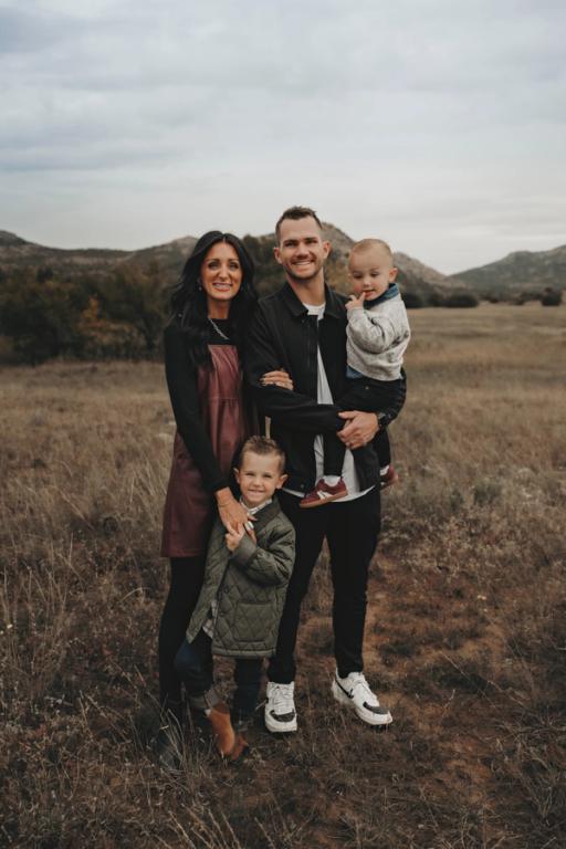 A smiling family of four, with two parents and two young sons, stands in a dry grassy field with distant mountains under an overcast sky.