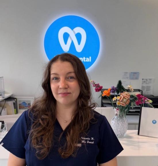 A woman with long, dark hair in blue scrubs smiles in a dental office, with a blue circular Win Dental sign in the background.