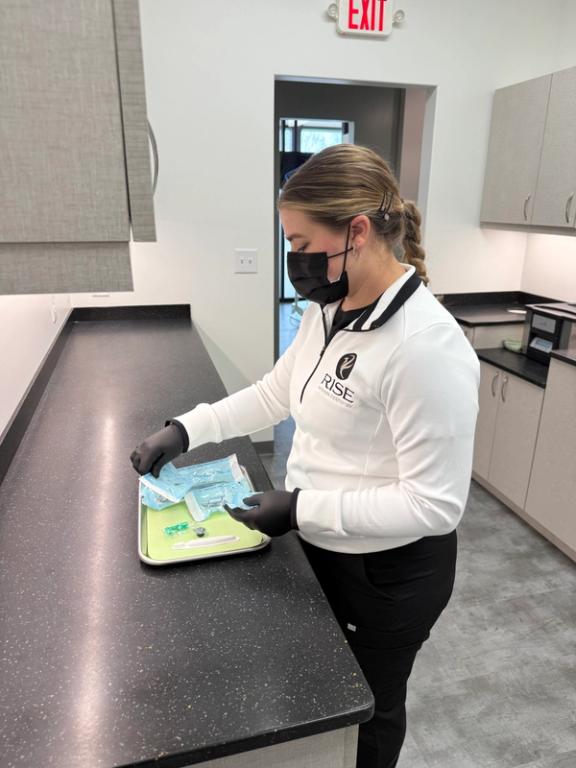A woman in a white uniform, black mask, and gloves prepares medical instruments on a tray.