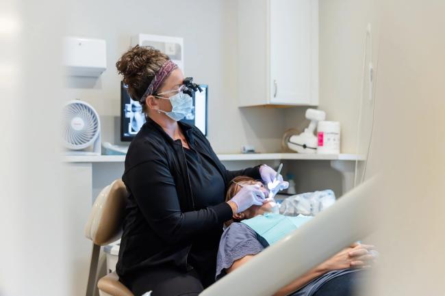 A masked dental professional with loupes examines a patient in a dental chair.