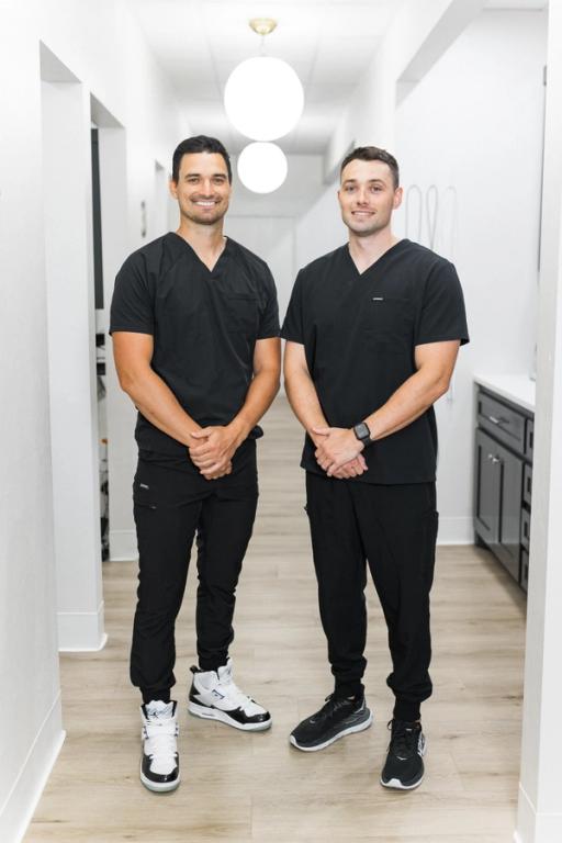 Two smiling men in black scrubs stand together in a modern hallway.