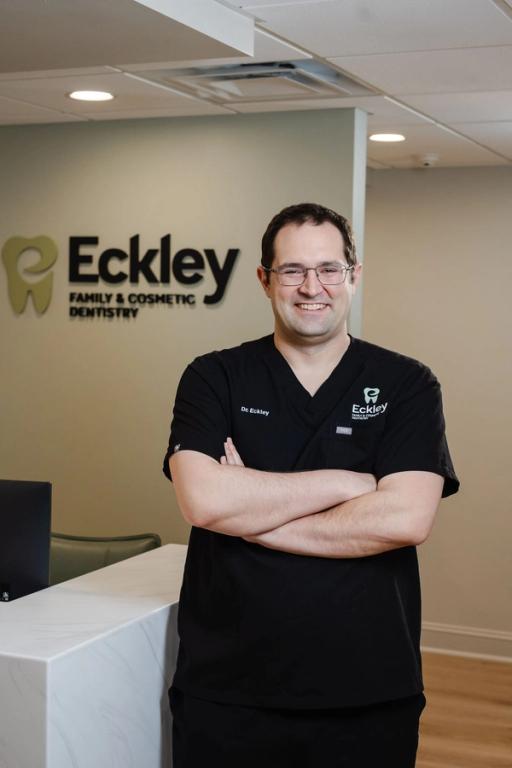 A smiling male dentist in black scrubs with crossed arms stands in front of the "Eckley Family & Cosmetic Dentistry" logo.