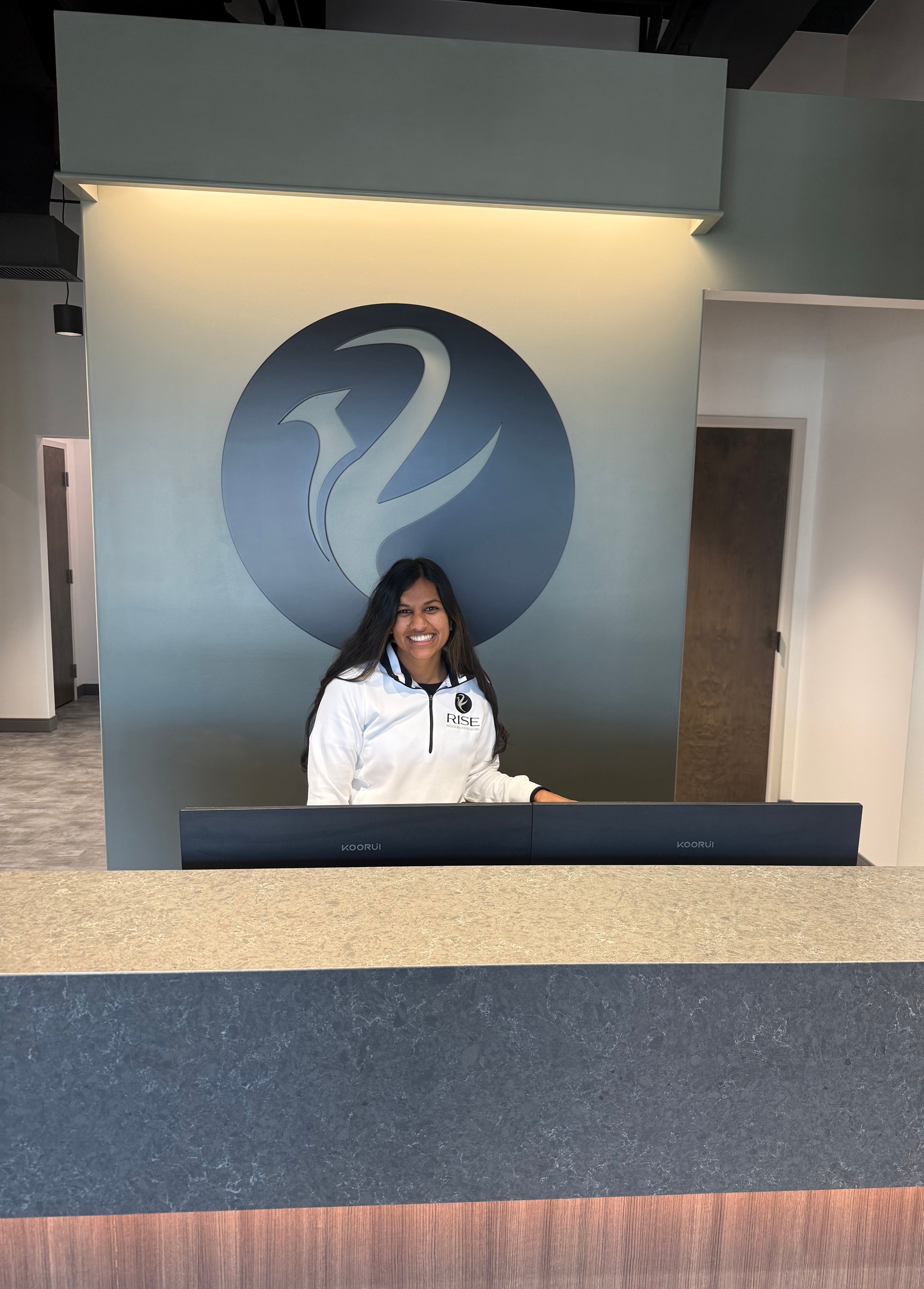 A smiling woman in a white jacket stands behind a reception desk with a large logo on the wall.