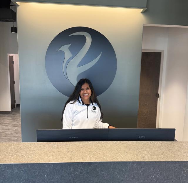 A smiling woman in a white jacket stands behind a reception desk with a large logo on the wall.